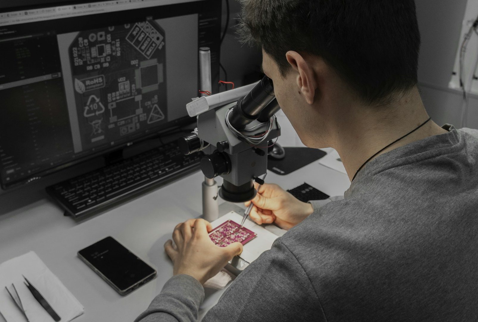 Sichtkontrollplatz mit Mikroskop Person arbeitet am Mikroskop an einer Platine, Computerbildschirm im Hintergrund.